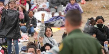 Las mujeres sostienen sus pulseras en alto con la esperanza de que los agentes de la Patrulla Fronteriza de EE. UU. las elijan para ser procesadas después de esperar días entre dos muros fronterizos.AFP